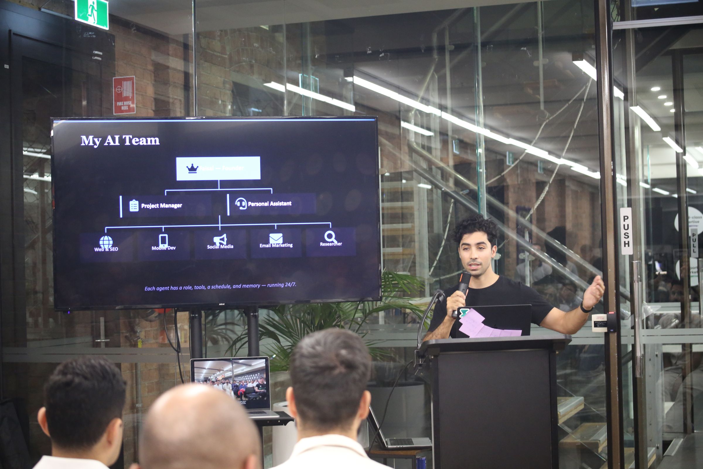Faisal Taibi at a lectern with a microphone, pointing to a slide titled “My AI Team” that shows an org chart of AI agents.